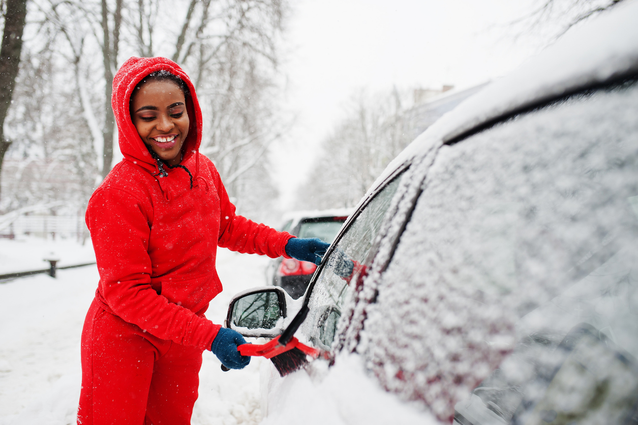 African american woman in red hoodie clean car from snow in winter day.