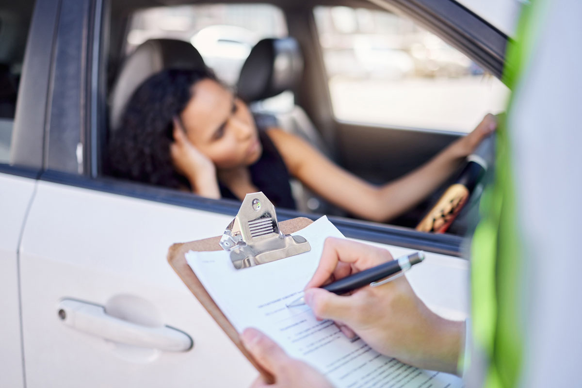A woman appears frustrated, sitting in her car while an officer fills out an accident report.