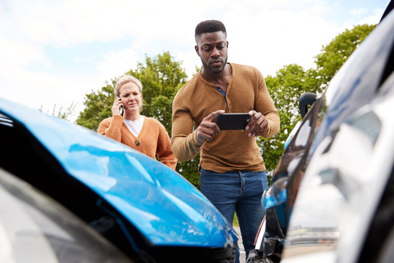A man takes photos of a car accident on his smart phone while the other drive looks on behind him.