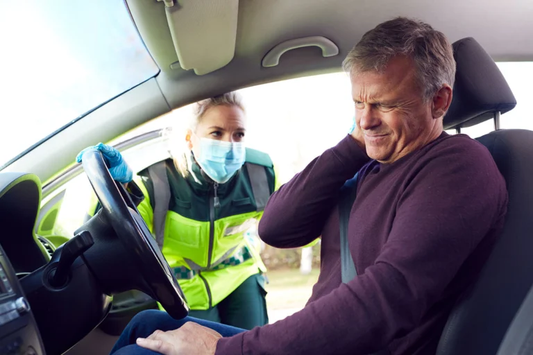A man sits behind the steering wheel of his car holding his neck just after an accident. A police officer stands outside checking on the man.
