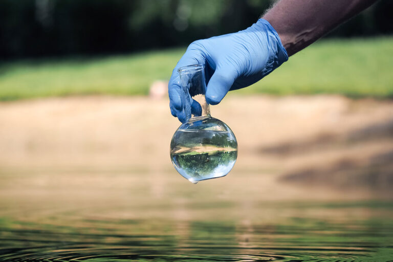 A person in blue gloves is holding a glass of water for PFAS analysis, showcasing the water sample collection process.