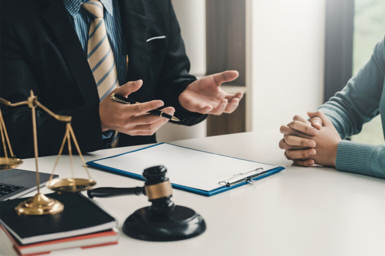 A lawyer and his clients sit at a table and discuss case related matters. A gavel and scales of justice are pictured on the table.