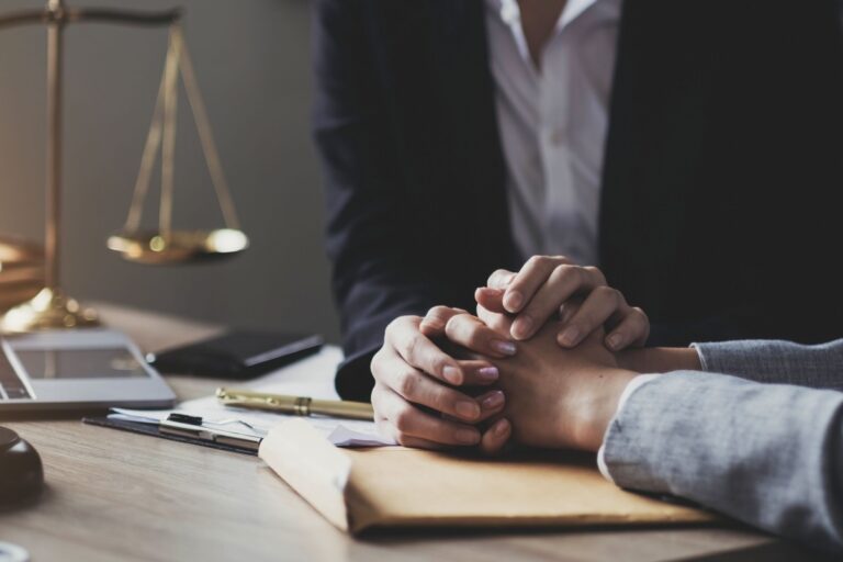 Two hands clasped on a desk with papers and a scale in the background, conveying support and empathy in a legal professional setting.