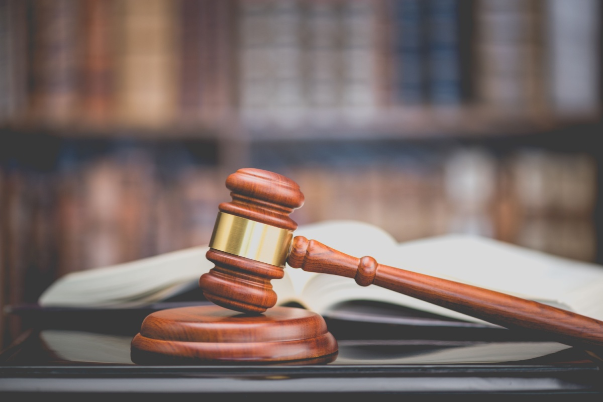Close-up of a wooden gavel on a table, with a bookshelf lined with books behind it.