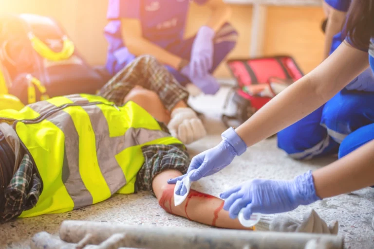 An individual wearing a blue vest and gloves is aiding a person on the ground, offering assistance in a critical workplace injury situation.
