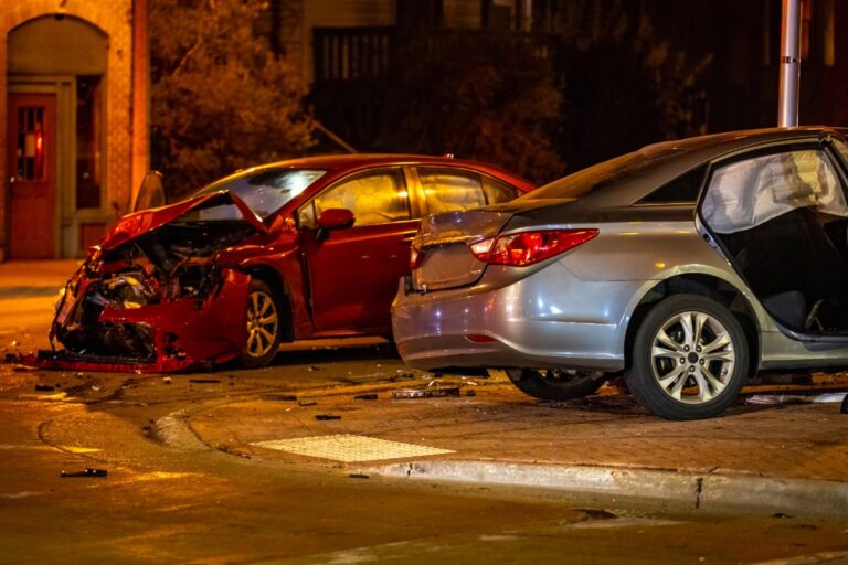 Two heavily damaged sedans after a nighttime collision—one red with massive front-end destruction, one silver with doors open—surrounded by road debris in an urban setting.