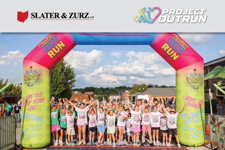 Group of smiling children celebrating at a Project Outrun Finish Line Festival under a large inflatable arch, proudly wearing custom-designed “I’m not sick, but my shoes are” t-shirts and shoes, with Slater & Zurz and Project Outrun logos visible.